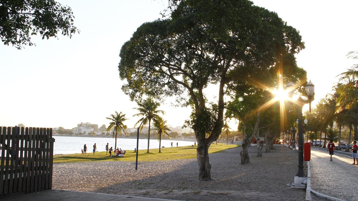 Orla da Lagoa de Araruama ao fim da tarde, com árvores, calçadão e o sol entre os galhos